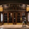 A young couple standing at the lit entrance of the Capitol Theatre at night, looking at the upcoming shows.