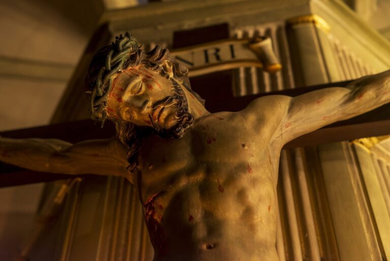 Painted wooden crucifix lit by warm candlelight in a church, seen from below.