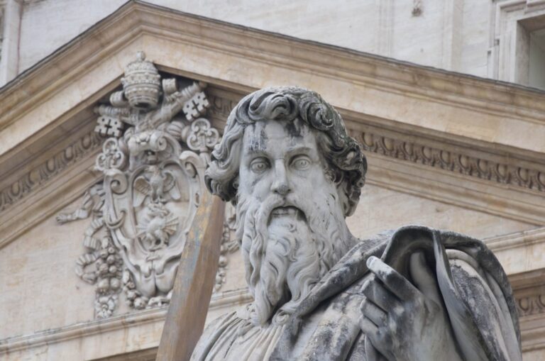Statue of St. Paul with his sword in front of the carved stone facade of St. Peter’s Basilica in Rome.